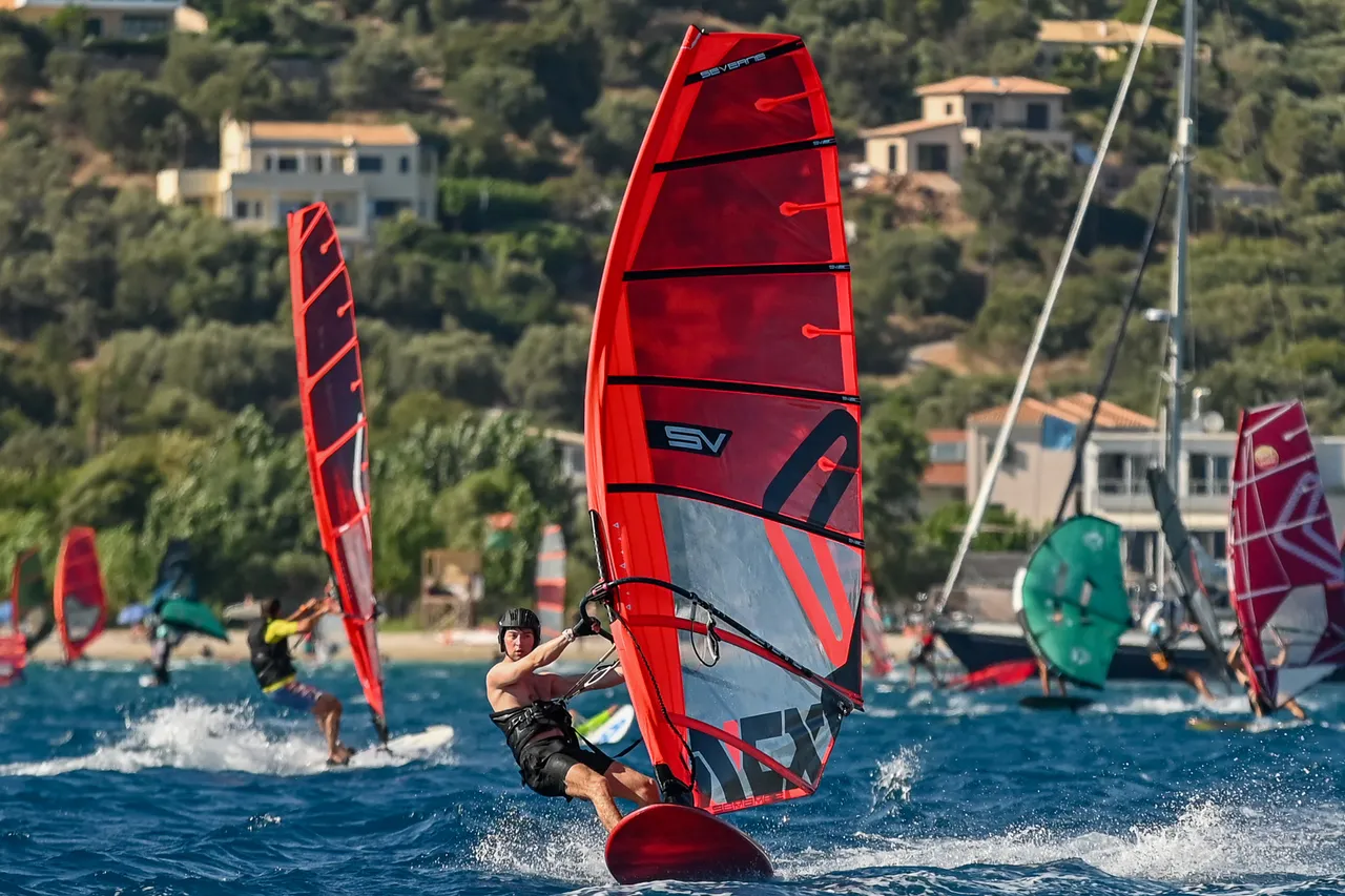 Ronan leaning back in the harness on a red windsurfer, sailing towards the camera with the beach and villas of Vassiliki behind.