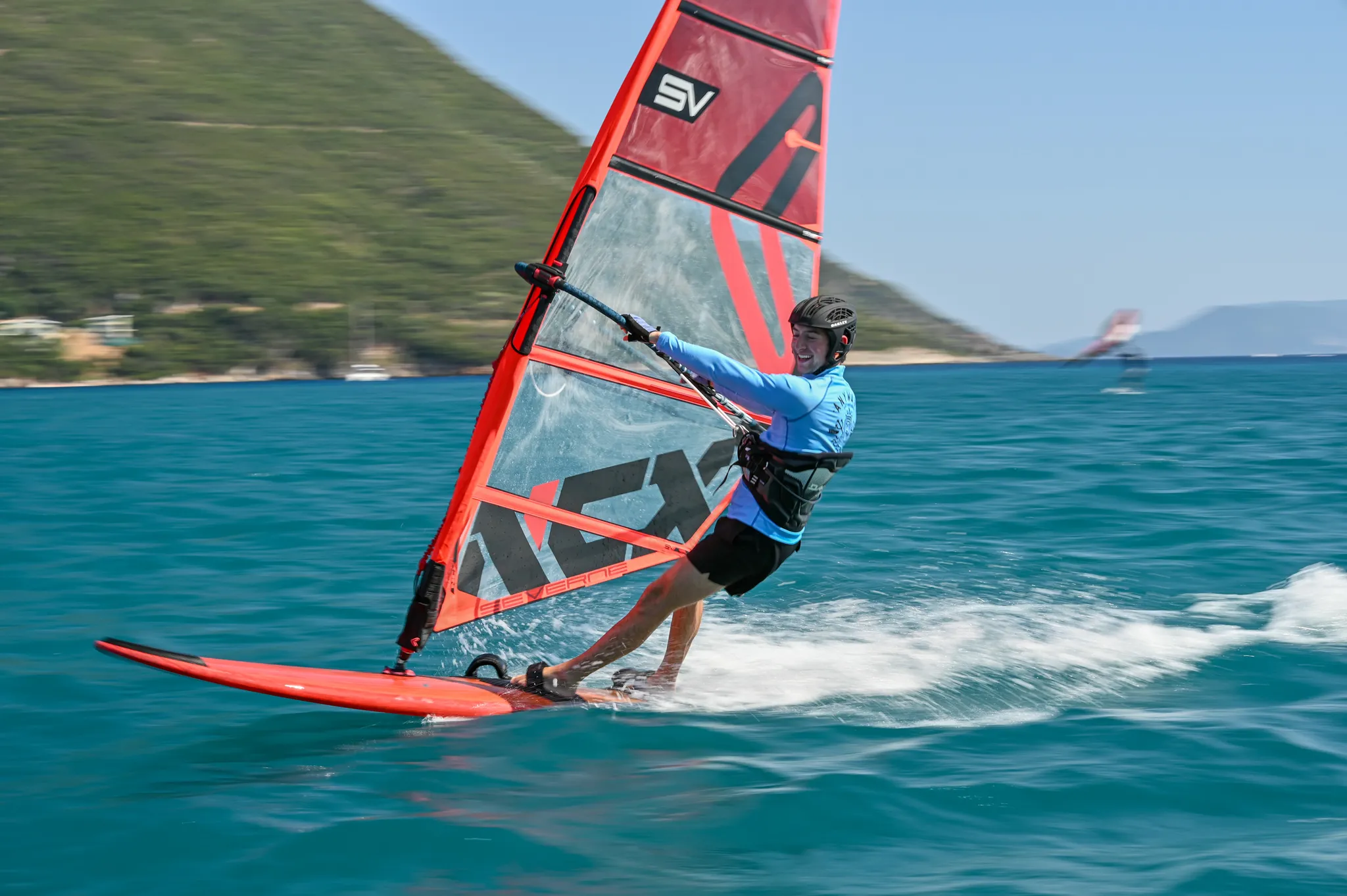 Ronan windsurfing at speed on a red board with a red and clear sail, grinning, with a green hillside behind him.